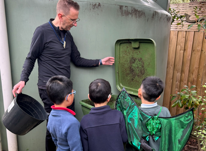 Teacher demonstrating composting to kids standing in front of a large green compost bin outdoors