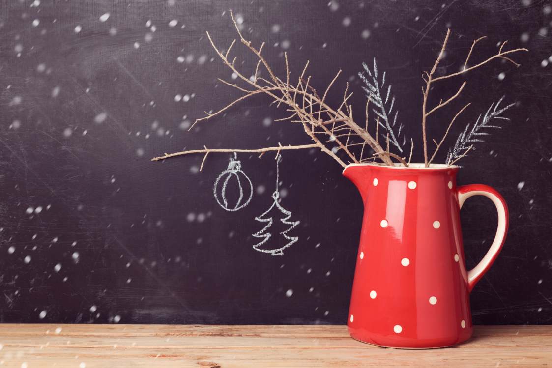 A red jug with white spots holds some small conifer branches with festive decorations.