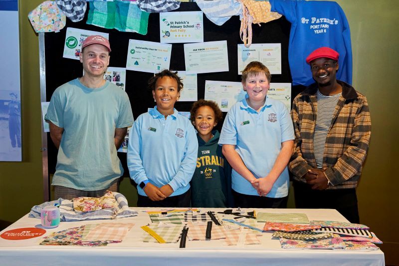 5 people standing behind a table displaying various handmade fabric items, including bags and pouches, at a community or school event.