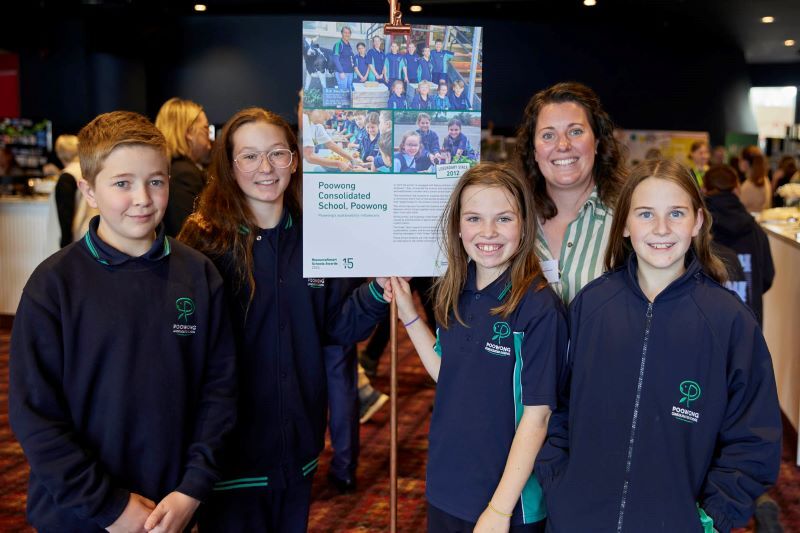 4r students and 1 adult standing indoors at an event, holding a sign labeled Poowong Consolidated School