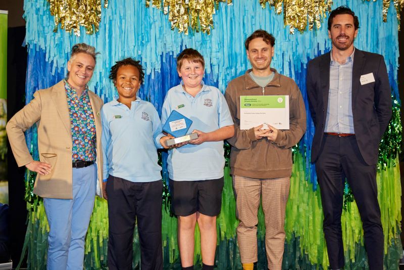 5 people standing in front of a colourful backdrop with blue, green, and gold streamers, posing for a group photo.
