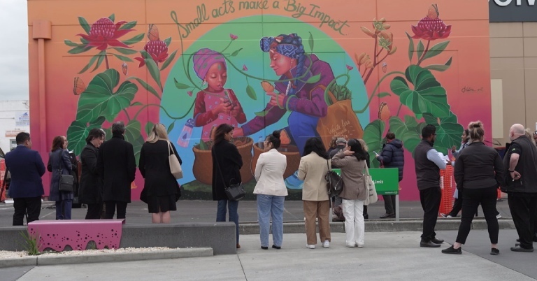 Group of people gathered outdoors in front of a colourful mural in St Albans depicting children planting a seedling, surrounded by large flowers and leaves