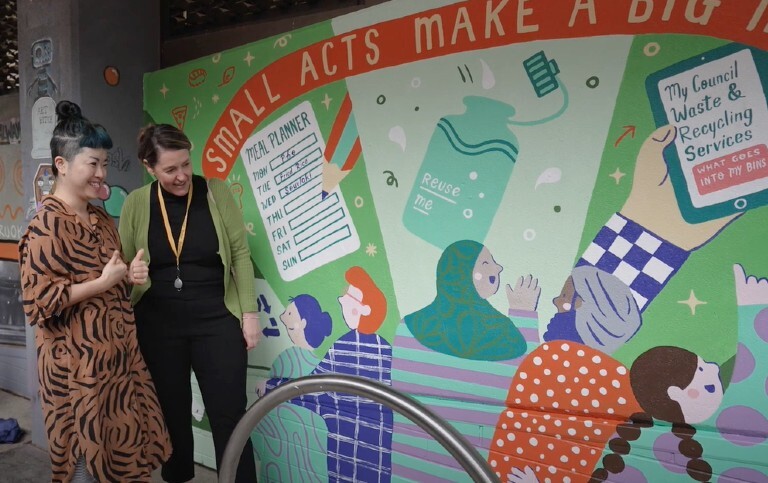 2 women standing next to a colourful mural promoting waste reduction and recycling, featuring illustrations of people, a reusable bottle, and a weekly meal planner